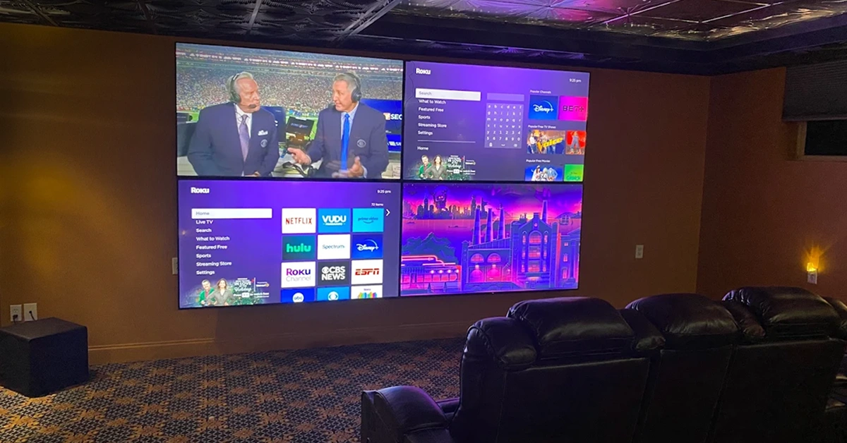 A professional installer from IhangTVs mounting a large flat-screen television onto a wall as part of a multi-screen video wall setup in a residential living room.