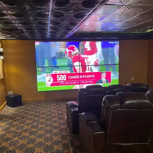 A professional home theater installation featuring a four-screen video wall configured to display a single large high-definition image of a college football game, set in a dedicated media room with a decorative tin ceiling, warm ambient lighting, and luxury black leather theater recliners.