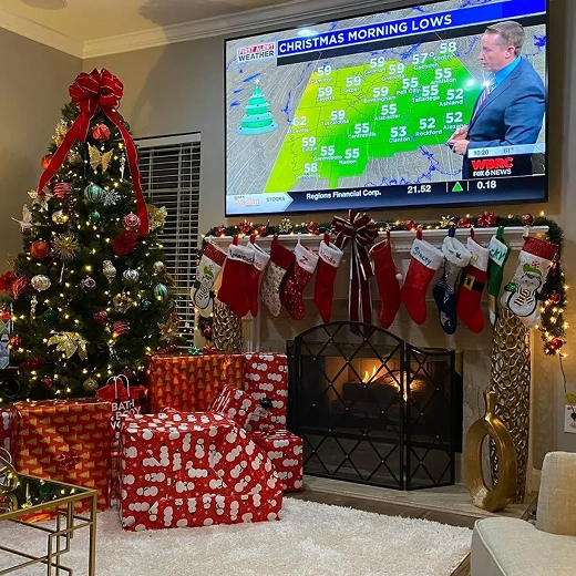 A professional holiday-themed TV installation featuring a large flat-screen television mounted above a white fireplace mantel. The festive living room is decorated with a tall Christmas tree, numerous wrapped gifts, and stockings hanging from a lit garland on the mantel, while the screen displays a local weather broadcast.