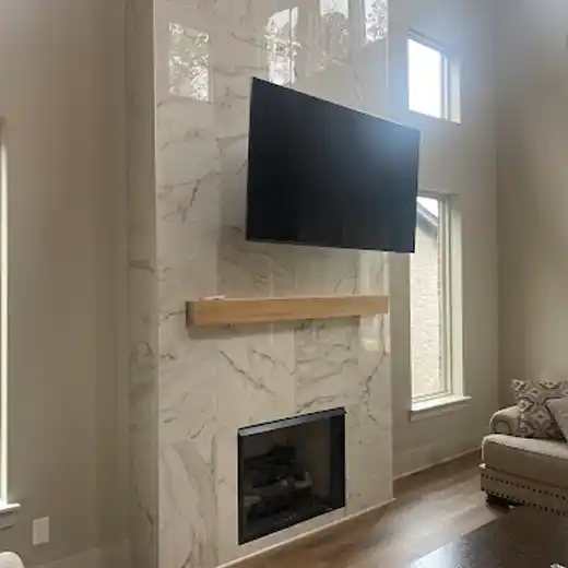 A professional TV installation in a luxury living room featuring a large flat-screen television mounted high on a floor-to-ceiling white marble-tiled fireplace wall, accented with a natural wood mantel and surrounded by tall windows that fill the space with natural light.