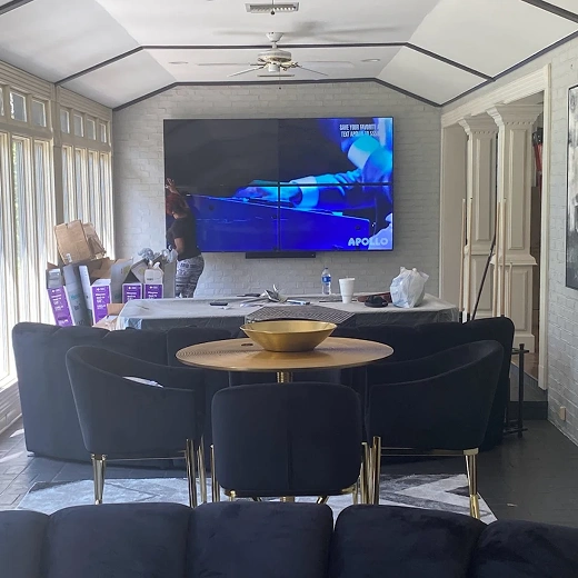 A professional video wall installation in a modern sunroom featuring four flat-screen monitors mounted together as a single large display on a white textured brick wall.