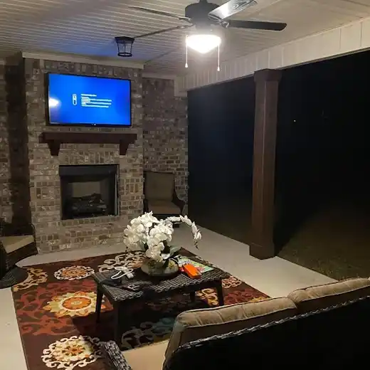A professional outdoor TV installation showing a large flat-screen television mounted above a brick fireplace on a covered patio, featuring comfortable wicker seating, a decorative area rug, and a ceiling fan with integrated lighting for an enhanced outdoor living space.
