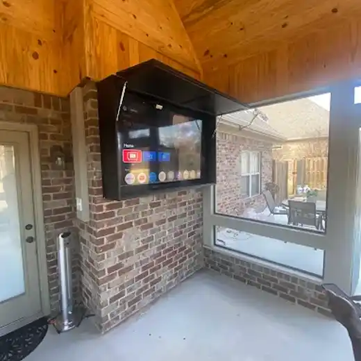 A professional outdoor TV installation on a screened-in porch showing a flat-screen television mounted inside a custom weather-protective black flip-up cabinet on a red brick pillar, set against high vaulted wood ceilings and large windows overlooking a backyard.