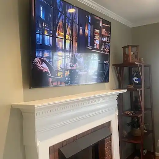 A professional TV installation showing a large flat-screen television mounted flush against a light grey wall above a traditional red brick fireplace with an ornate white mantel, displaying a high-definition image of a cozy indoor cafe scene.