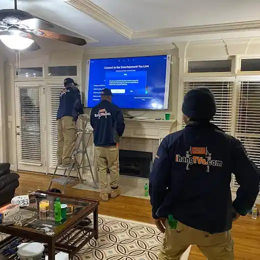 A professional TV mounting installation by the ihangTVs team in a living room, showing three technicians in branded uniforms setting up a large flat-screen television over a fireplace mantel while a digital setup screen is displayed.