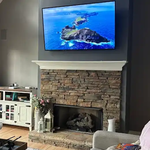 A professional TV installation showing a large flat-screen television mounted securely on a dark grey accent wall above a rustic stone fireplace with a white mantel, featuring a clean look with no visible wires in a modern living room setting.
