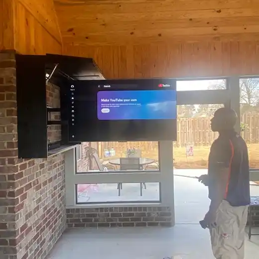 A professional TV installation in an enclosed sunroom showing a flat-screen television mounted with a specialized swivel arm inside a custom black cabinet on a brick wall, featuring a wood-paneled ceiling and a technician finalizing the setup.